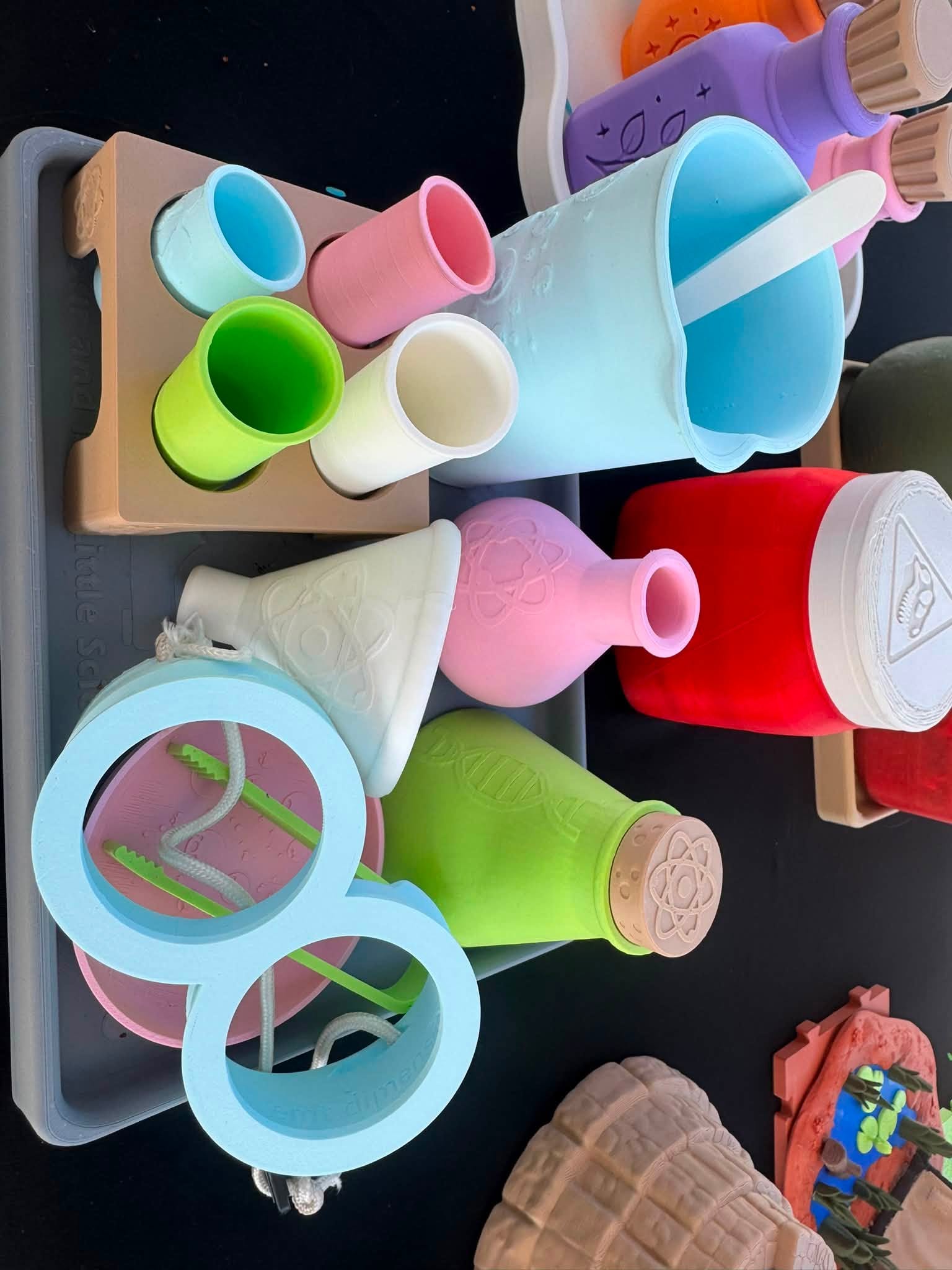 Colourful 3D-printed Little Scientist prototypes including flasks, cups, funnels and play goggles arranged on a workshop table.